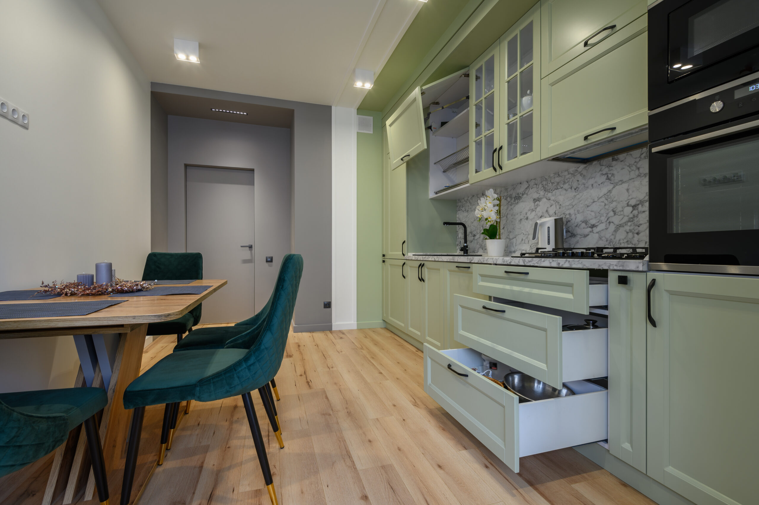 Modern trendy light green luxurious kitchen with dining table and dark green chairs, some drawers are retracted