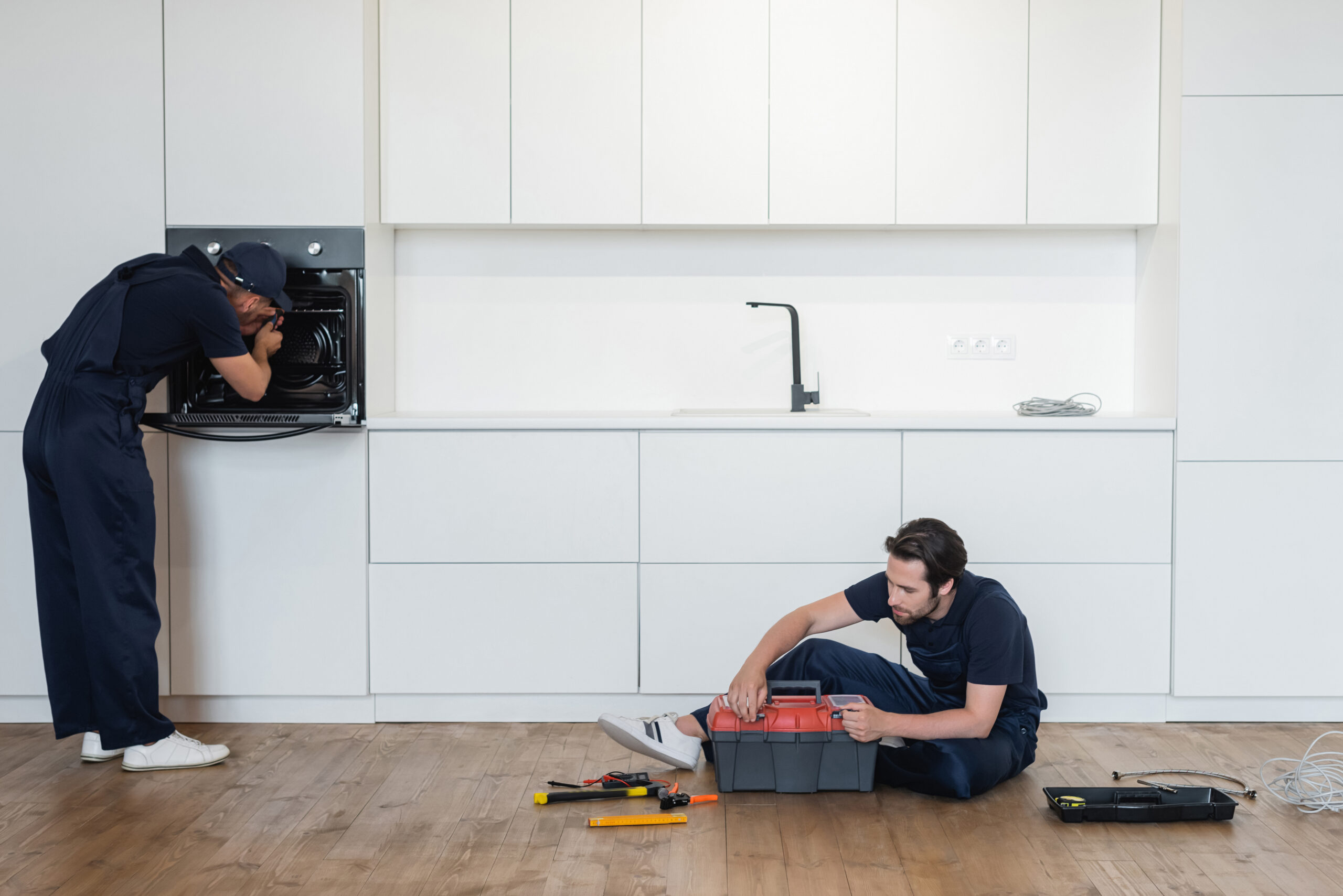 repairman fixing electric oven near colleague sitting on kitchen floor with toolbox
