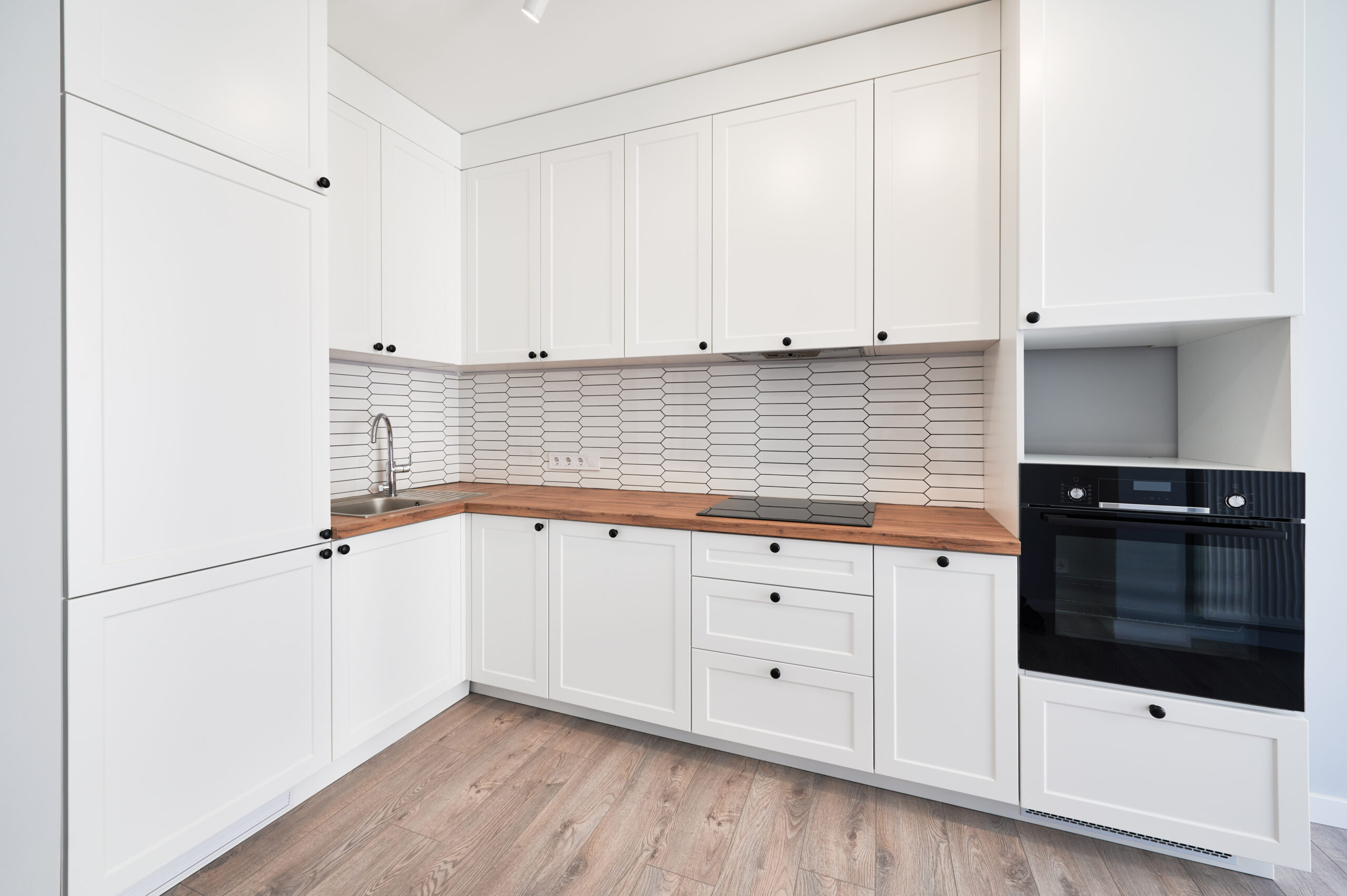 Kitchen with laminate floor, white walls, doorway and modern kitchen set with electric stove, counter, sink and oven. Apartment kitchen room with elegant minimalist design in white colors.