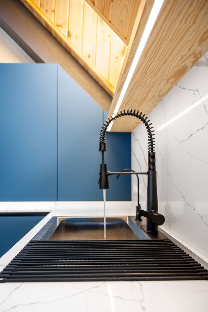Vertical view of a professional spring faucet with a steady water stream, roll up drainer and veined marble. Blue handleless fronts and a slim LED line deliver a clean, minimalist look.