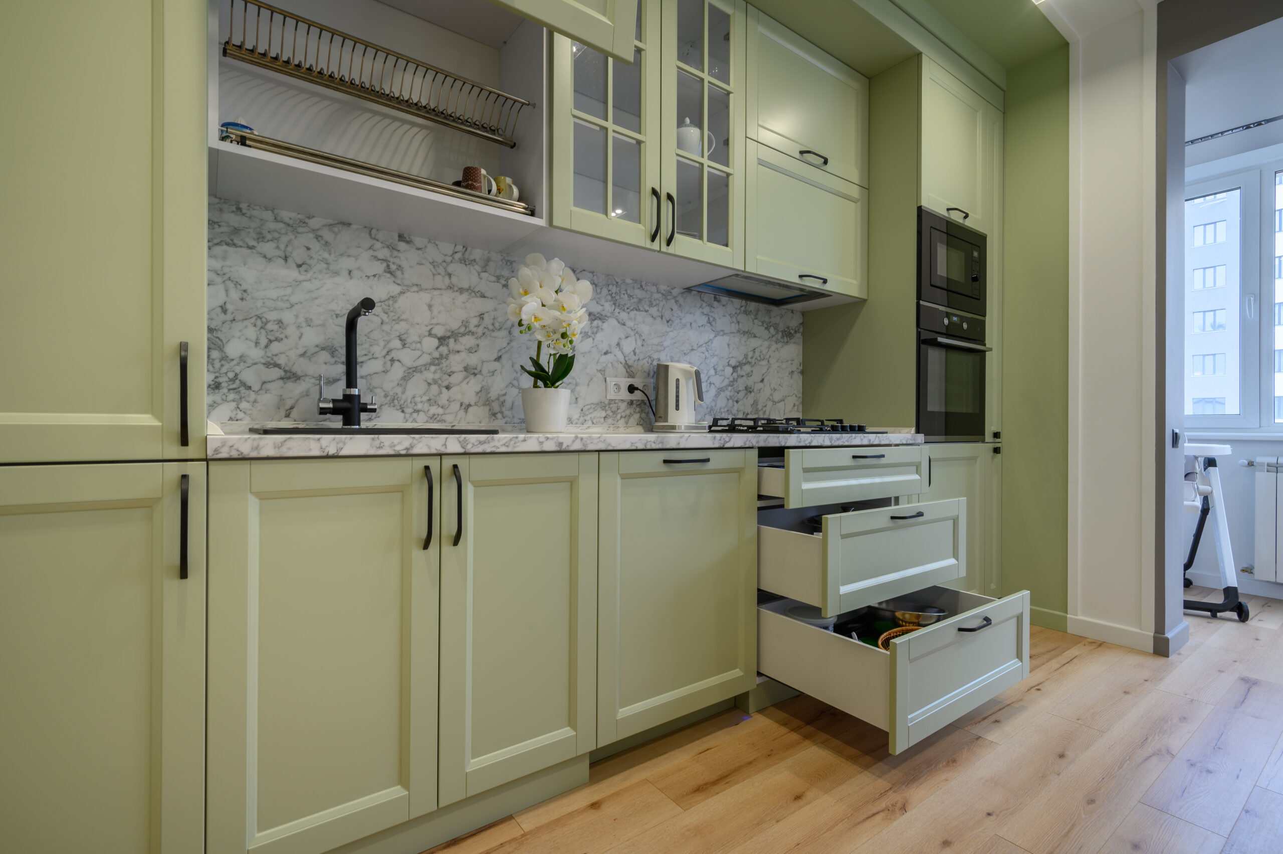 Modern trendy light green luxurious kitchen with dining table and dark green chairs, some drawers are retracted