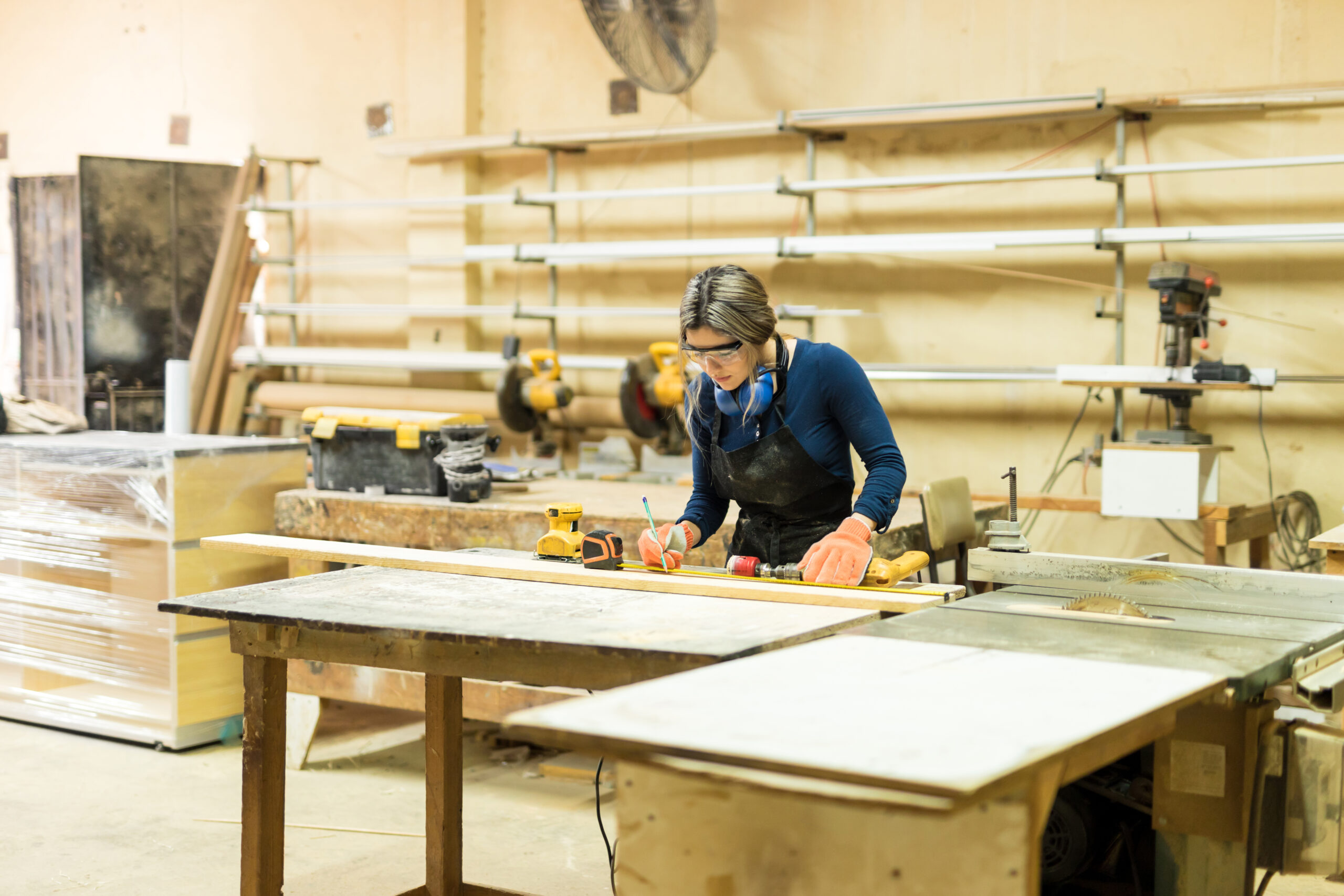 Wide view of a busy female carpenter measuring and marking some wood in her workshop