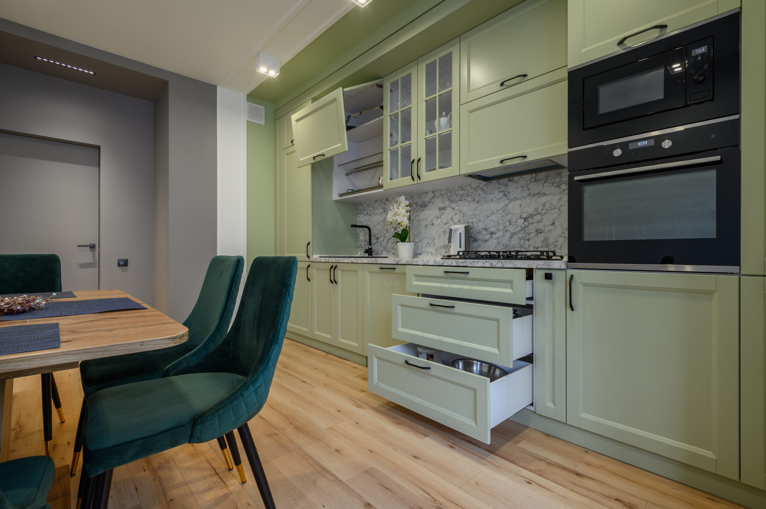 Modern trendy light green luxurious kitchen with dining table and dark green chairs, some drawers are retracted